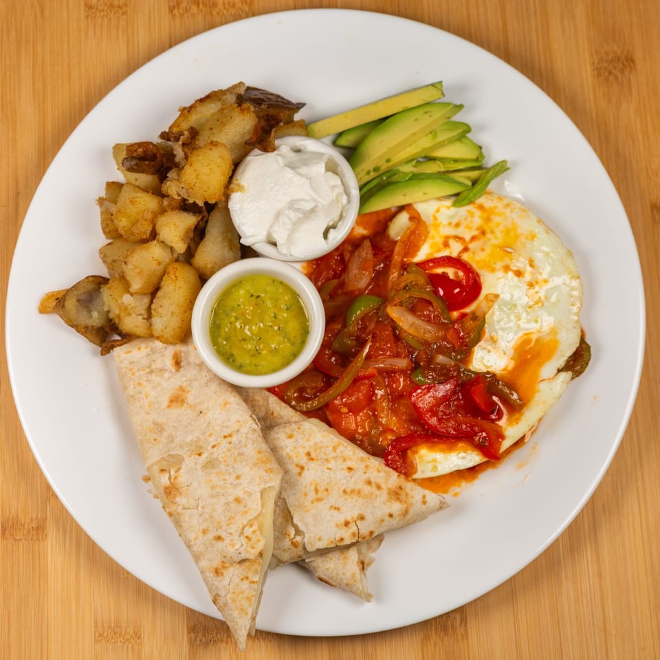 Breakfast plate with fried eggs, bell peppers, onions, potatoes, avocado, sour cream, and salsa.